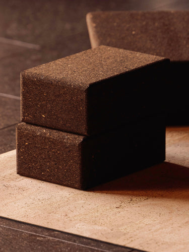 Three dark brown textured cork yoga blocks stacked and standing on a beige mat over a dark tiled floor.