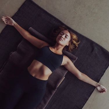 Woman lying on a natural non slip cotton yoga rug in a restorative yoga pose. #color_ash