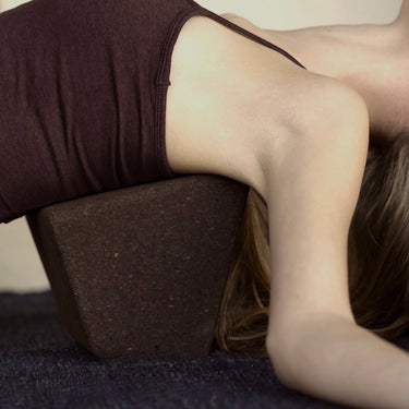 woman laying on earth or brown cork restorative yoga block on top of ash or black yoga rug