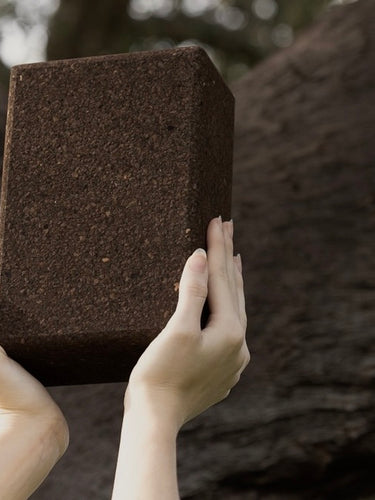 woman holding a natural rituil cork yoga block in front of a tree
