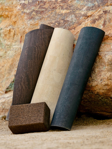 Rituil cream and black cork leather yoga mats featuring a non-slip, non-toxic, pvc free surface rolled up and leaning on rock.