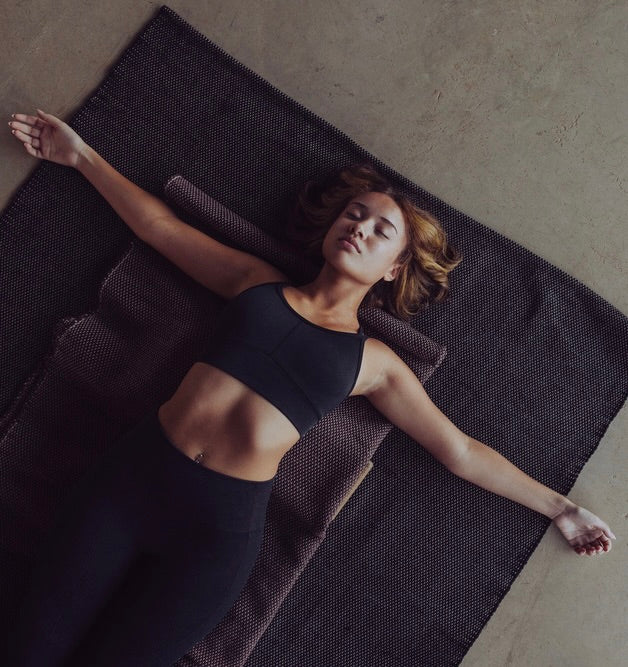 Woman lying on a natural non slip cotton yoga rug in a restorative yoga pose. #color_ash